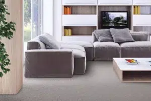Modern living room with a grey sectional sofa, a low wooden coffee table, and white shelving with a mounted TV. A large window and greenery are visible on the left.