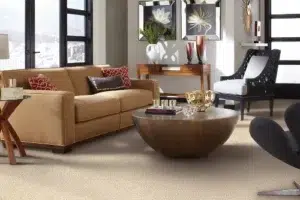 Modern living room with a tan leather sofa, red patterned and black pillows, a large round coffee table, and a beige carpet; black-and-white armchair near large windows, with floral wall art and a wooden console against the wall.
